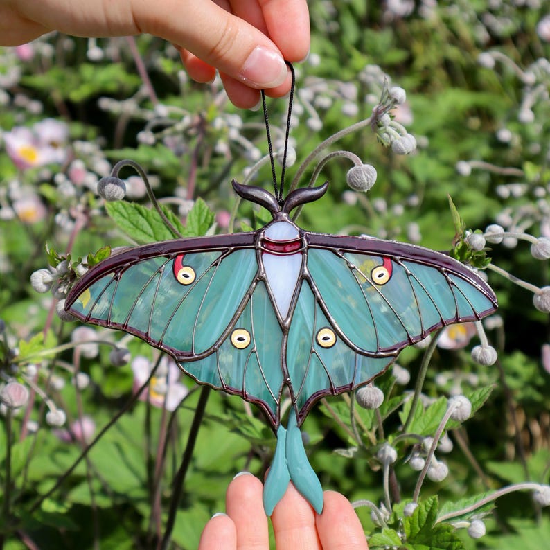 May include: A stained glass luna moth suncatcher with teal wings, a white and red body, and a dark purple border. The moth has yellow and red accents and is suspended by a black string. The background is a blurred view of green foliage and flowers.