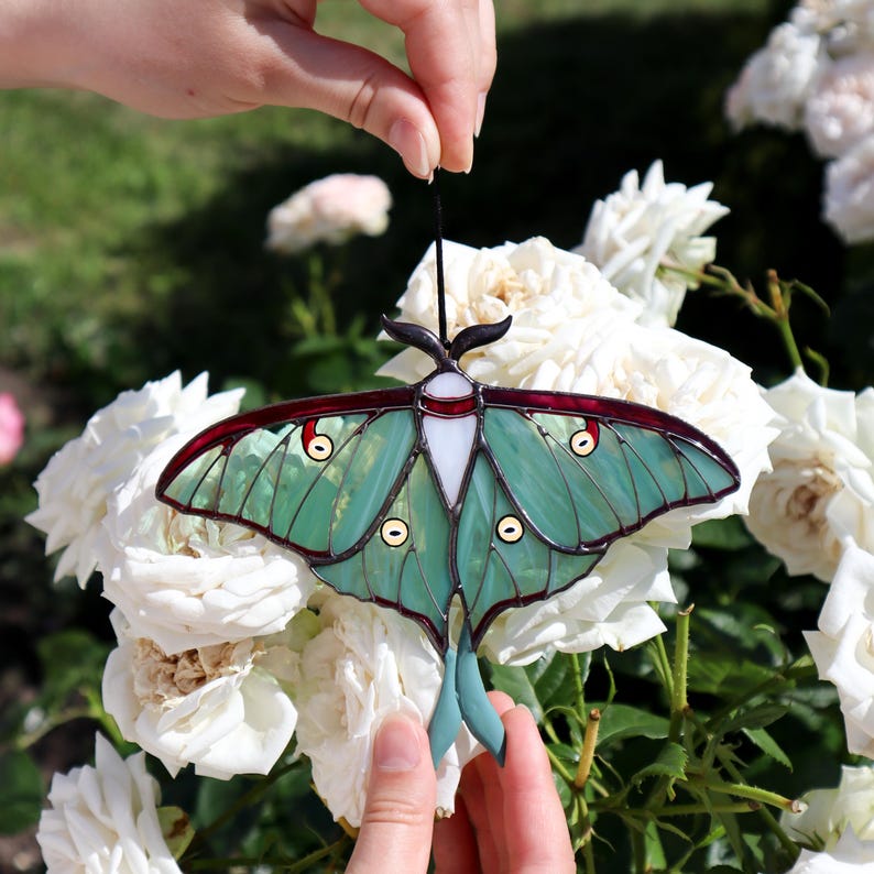 May include: A stained glass Luna moth suncatcher with a teal and red wing design. The moth has a white body and yellow and black eye spots. The suncatcher is held up by a black string against a background of white roses.