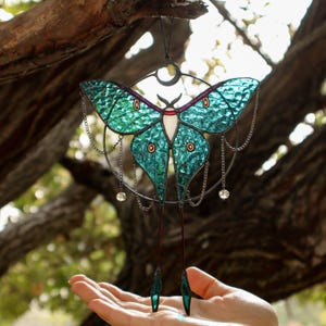May include: A stained glass moon moth suncatcher with a teal and green color scheme. The moth is surrounded by a circle of chains with crystal beads hanging from them.