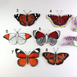 May include: A collection of colorful butterfly ornaments with intricate designs. The butterflies feature various patterns and colors, including red, orange, white, and black. Each butterfly has a unique wing pattern, with details like black spots and lines.