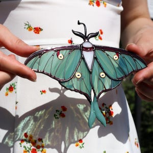 May include: A stained glass luna moth with teal wings, a white body, and dark antennae. The wings feature circular yellow accents and are outlined in a dark metal. The moth is held against a white floral background.