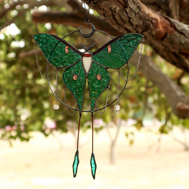 May include: A stained glass moon moth hanging from a branch. The moth is green with red and white accents. The moth is surrounded by a circle of metal with chains and beads hanging from it.