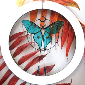May include: A teal blue stained glass butterfly with black and silver accents hanging from a silver chain. The butterfly is suspended in a white circle with a white background. The butterfly has a crescent moon shape at the top of its body.
