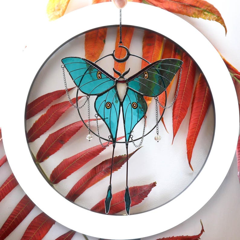 May include: A teal blue stained glass butterfly with black and silver accents hanging from a silver chain. The butterfly is suspended in a white circle with a white background. The butterfly has a crescent moon shape at the top of its body.