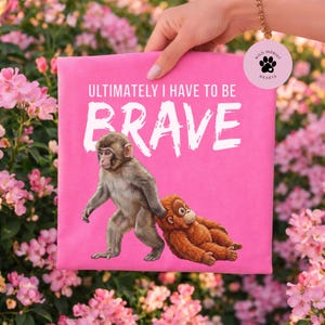 Op de afbeelding: Een roze t-shirt met de witte tekst "ULTIMATELY I HAVE TO BE BRAVE". Het shirt heeft een afbeelding van twee apen, één lopend en de ander liggend. Het shirt heeft ook een "Wild Imprint Hearts" logo.