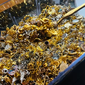 May include: A clear container overflowing with an assortment of gold and silver-toned jewelry pieces. The collection includes various styles of earrings, pendants, and charms. A gold-colored spoon rests on top of the jewelry.