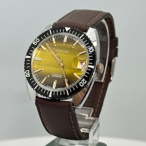 May include: A gold-toned wristwatch with a black bezel and a brown leather strap. The watch face has a yellow dial with black markings and the text "VIALUX 17 JEWELS INCABLOC SWISS MADE".