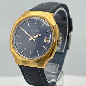 May include: A gold-tone wristwatch with a blue face and a black leather strap. The watch face has white markings and the word "VIALUX" printed on it. The watch has a date window at the 3 o'clock position.