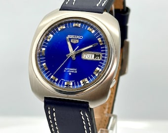 May include: A silver Seiko automatic watch with a blue face and a black leather strap. The watch face has a date window that says "TUE".