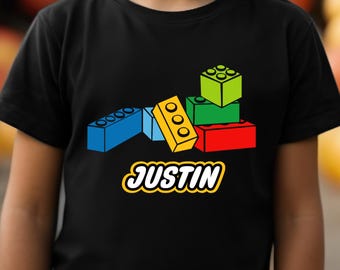 May include: Black t-shirt featuring a colorful graphic of toy building blocks in blue, yellow, green, and red. The name "JUSTIN" is printed in a yellow and white font below the blocks. The shirt is made of a soft material.