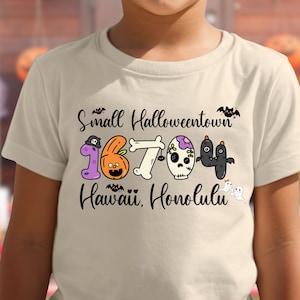 May include: A beige t-shirt featuring the text "Small Halloweentown" above a graphic of the numbers "16704" with Halloween-themed designs. Below the numbers is the text "Hawaii Honolulu".