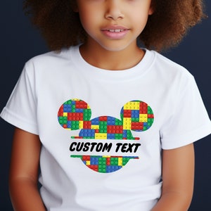 May include: White t-shirt with a colorful Mickey Mouse head design made of Lego bricks. The design includes the text "CUSTOM TEXT" underneath the head.