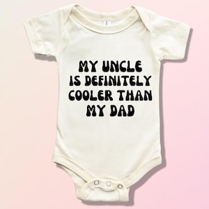 May include: White baby onesie with black text that reads "My Uncle is Definitely Cooler Than My Dad".