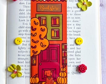 May include: A clear bookmark featuring a bookstore illustration with a red door, pumpkins, and a dog. The bookmark has the word "Bookstore" at the top. The bookmark is placed in an open book with floral embellishments.