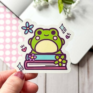 May include: A green frog with a yellow belly sitting on top of a stack of three colorful books. The books have flowers on them and there are sparkles and hearts around the frog.