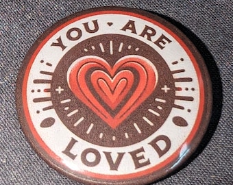 Pode incluir: Um botão redondo com fundo marrom e borda branca. O botão tem um coração vermelho com círculos concêntricos no centro. O texto "YOU ARE LOVED" está escrito em branco ao redor do coração.