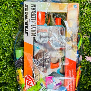 Pu&ograve; includere: Un cesto blu intrecciato pieno di un set di giocattoli di Star Wars: The Mandalorian, caramelle e un fiocco blu. Il cesto &egrave; avvolto in cellophane trasparente. La scatola dei giocattoli &egrave; arancione e bianca.