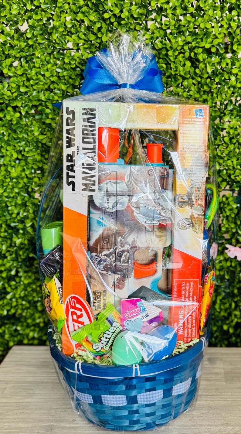 Pu&ograve; includere: Un cesto blu intrecciato pieno di un set di giocattoli di Star Wars: The Mandalorian, caramelle e un fiocco blu. Il cesto &egrave; avvolto in cellophane trasparente. La scatola dei giocattoli &egrave; arancione e bianca.