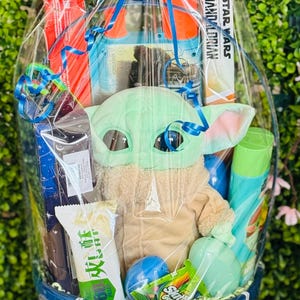 Pu&ograve; includere: Un cesto regalo blu pieno di vari articoli, tra cui un peluche di Grogu, un libro di Star Wars Mandalorian e snack. Il cesto &egrave; avvolto in cellophane trasparente e legato con un grande fiocco blu. Contiene anche prodotti per il bagno e caramelle.