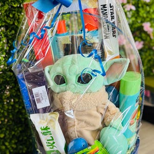 Pu&ograve; includere: Un cesto regalo in vimini blu e bianco avvolto in cellophane trasparente e legato con un grande fiocco blu. Il cesto contiene un peluche di Grogu, un libro di Star Wars Mandalorian e vari altri oggetti.