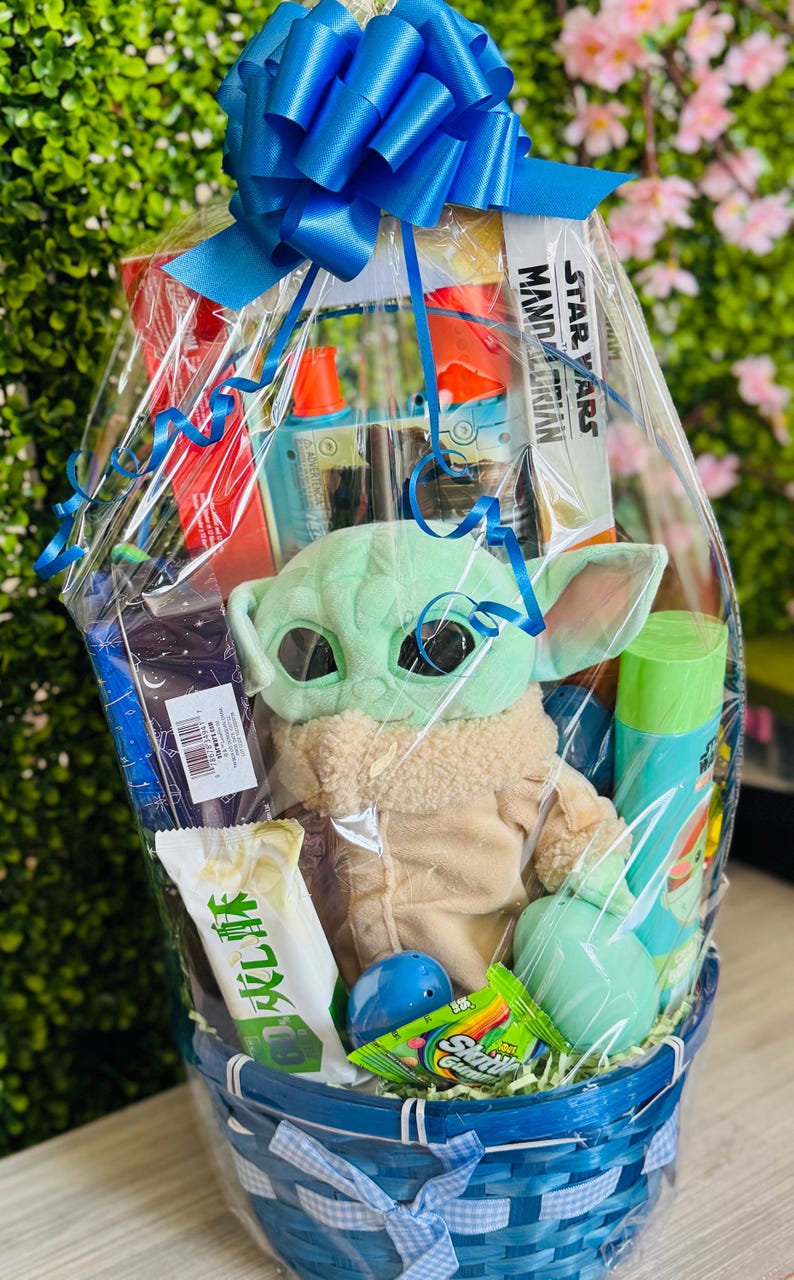 Pu&ograve; includere: Un cesto regalo in vimini blu e bianco avvolto in cellophane trasparente e legato con un grande fiocco blu. Il cesto contiene un peluche di Grogu, un libro di Star Wars Mandalorian e vari altri oggetti.