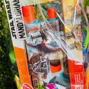 Pu&ograve; includere: Un cesto regalo con un giocattolo di Star Wars: The Mandalorian, caramelle e uova di Pasqua, avvolto in cellophane trasparente e legato con un nastro blu. Il cesto include un blaster giocattolo con dettagli arancioni.
