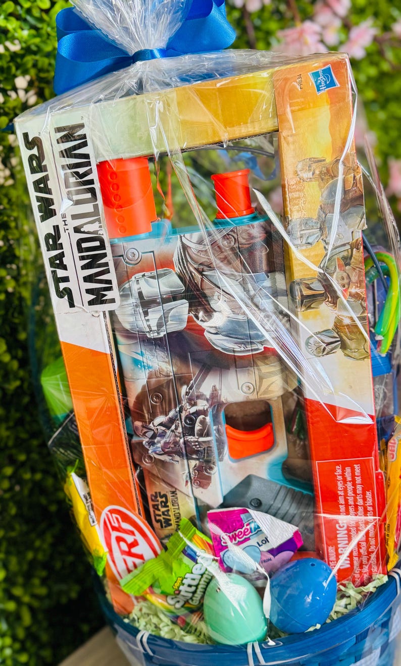 Pu&ograve; includere: Un cesto regalo con un giocattolo di Star Wars: The Mandalorian, caramelle e uova di Pasqua, avvolto in cellophane trasparente e legato con un nastro blu. Il cesto include un blaster giocattolo con dettagli arancioni.