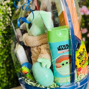Pu&ograve; includere: Un cesto blu intrecciato pieno di regali a tema Star Wars. Il cesto contiene un pupazzo di peluche di Grogu, una bottiglia di bagnoschiuma al profumo di agrumi e varie caramelle. Il cesto &egrave; avvolto in cellophane trasparente e legato con un nastro blu.
