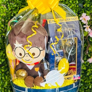 Könnte beinhalten: Ein blauer Korb gefüllt mit Harry Potter-Geschenken, darunter ein Plüschtier, Socken und Süßigkeiten. Der Korb ist in durchsichtiges Cellophan gehüllt und mit einer großen gelben Schleife versehen. Der Korb ist mit gelbem Krepppapier gefüllt.