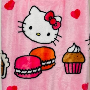 Könnte beinhalten: Rosa Plüschdecke mit Hello Kitty, Makronen, Cupcakes und Herzen. Die Decke hat ein sich wiederholendes Muster von Hello Kittys Gesicht, Schleifen und Desserts. Das Hello Kitty-Logo ist auf der Verpackung sichtbar.
