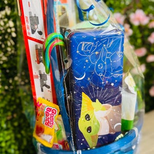 Pu&ograve; includere: Un cestino blu intrecciato pieno di regali, tra cui un astuccio blu con un design di personaggi dei cartoni animati, caramelle e una pistola giocattolo. Il cestino &egrave; avvolto in cellophane trasparente e legato con un nastro blu.
