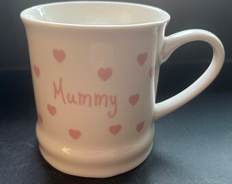 May include: A white ceramic mug with a curved handle. The mug is decorated with pink hearts and the word "Mummy" in pink script. The mug has a slightly flared rim and a rounded base.