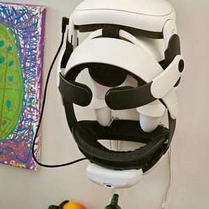 May include: A white virtual reality headset with black adjustable straps and a black facial interface cushion, mounted on a wall. The device features a white battery pack attached to the bottom and a black charging cable connected to the side.