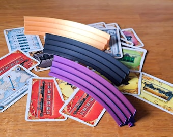 May include: Three curved, multi-grooved plastic track segments in orange, black, and purple are stacked on top of a scattered collection of rectangular board game cards featuring illustrations of trains, maps, and various game-related text.