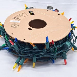 May include: A circular cardboard spool labeled PolyTerra PLA wrapped with a long strand of dark green electrical wire featuring multi-colored incandescent light bulbs in red, blue, yellow, and green, with a standard electrical plug attached.