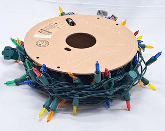May include: A circular cardboard spool labeled PolyTerra PLA wrapped with a long strand of dark green electrical wire featuring multi-colored incandescent light bulbs in red, blue, yellow, and green, with a standard electrical plug attached.