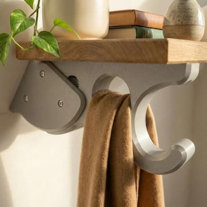 May include: A light gray wall-mounted hook with a curved design supporting a wooden shelf. A tan fabric item hangs from the hook. The shelf holds a potted plant, two books, a speckled ceramic vase, and a white candle.