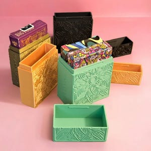 May include: A collection of rectangular playing card cases in various colors including mint green, yellow, black, and gold. Each case features an embossed floral pattern on the exterior. Some cases are open, while others hold decks of cards.