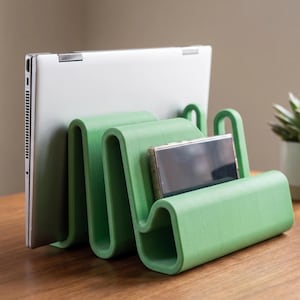 May include: A light green, wavy-shaped desk organizer with multiple slots. A silver laptop is positioned in the back slot, and a rectangular smartphone is placed in the front slot. The organizer has a smooth, matte finish and a continuous, folded design.
