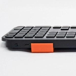 May include: A low-profile black wireless keyboard featuring a rectangular orange plastic riser attached to the side. The keyboard has a USB-C charging port and a small circular power button visible on the side edge near the orange support piece.