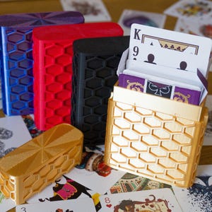 May include: A collection of five rectangular playing card cases featuring a raised honeycomb pattern on the exterior. The cases are colored in metallic blue, red, matte black, and two in metallic gold, with one gold case holding a deck of cards.