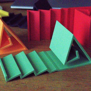 May include: A collection of 3D printed plastic stands in orange, green, and red. The green stand features a stepped, zig-zag base with a triangular support. The orange stand has a similar triangular design. A red stand with a tall, folded structure stands in the back.