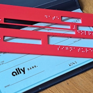 May include: A rectangular red plastic cheque writing guide featuring cutouts for writing and raised white braille dots along the edges. The guide is positioned over a light blue Ally Bank cheque, which includes the printed text "ally BANK" and "MEMO".