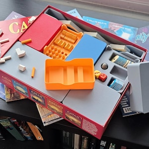 May include: A Catan board game box containing various plastic storage inserts in orange, blue, and gray. The box includes small wooden game pieces, two six-sided dice, and stacks of cards. The red box exterior features the Catan logo and artwork.
