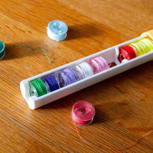 May include: A white plastic storage case containing multiple colorful sewing machine bobbins with thread in shades of green, blue, purple, white, pink, red, and yellow, with additional loose bobbins placed nearby on the surface.