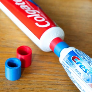 Pode incluir: Um tubo de pasta de dentes Colgate ligado a um tubo de pasta de dentes Crest através de um adaptador de plástico vermelho e azul. Dois adaptadores soltos adicionais, um vermelho e um azul, estão posicionados perto. Os tubos exibem os logótipos.