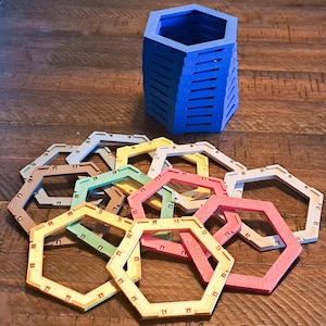 May include: A stack of blue hexagonal interlocking components sits behind a collection of flat, open-frame hexagonal rings in various colors including yellow, pink, green, tan, and white, all featuring small rectangular notches along their edges.