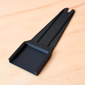May include: A black, tapered plastic tool featuring a flat, square-shaped base at one end and a narrow, elongated handle with a small U-shaped notch at the tip. The object has a matte finish and a rigid, structured design with visible ridges.