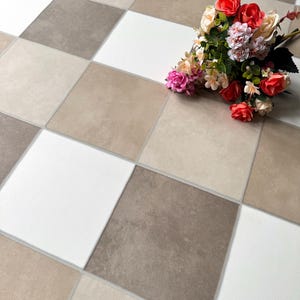 May include: A checkered floor pattern in shades of white, beige, and brown. A bouquet of colorful flowers with red, pink, and cream-colored blooms rests on the floor. The tiles are square and arranged in a grid.