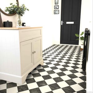 May include: A white cabinet with a light wood top stands on a floor featuring a black and white chequered pattern. The cabinet has two doors with silver knobs. A glass vase with green foliage sits on the surface next to a black container.